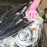 A person pours car wash soap into a bucket beside a hose and wash mitt