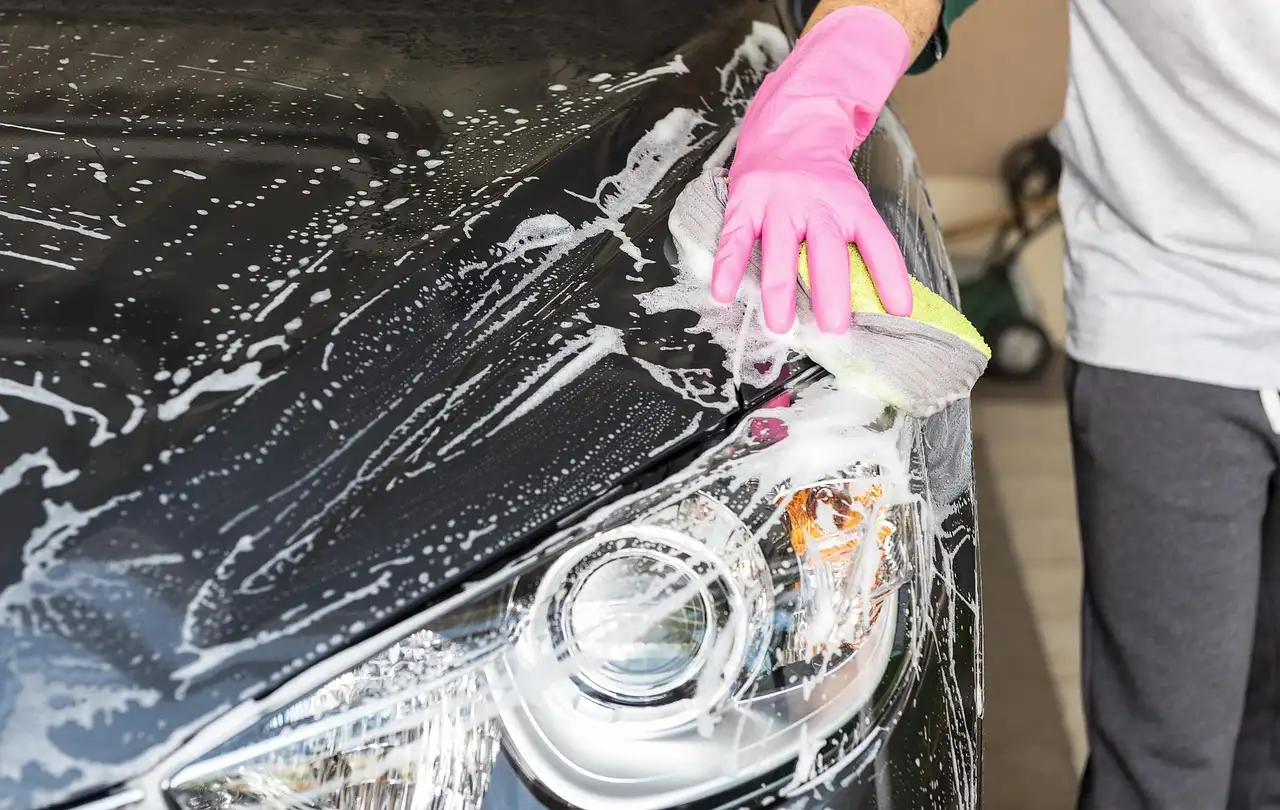 A person pours car wash soap into a bucket beside a hose and wash mitt